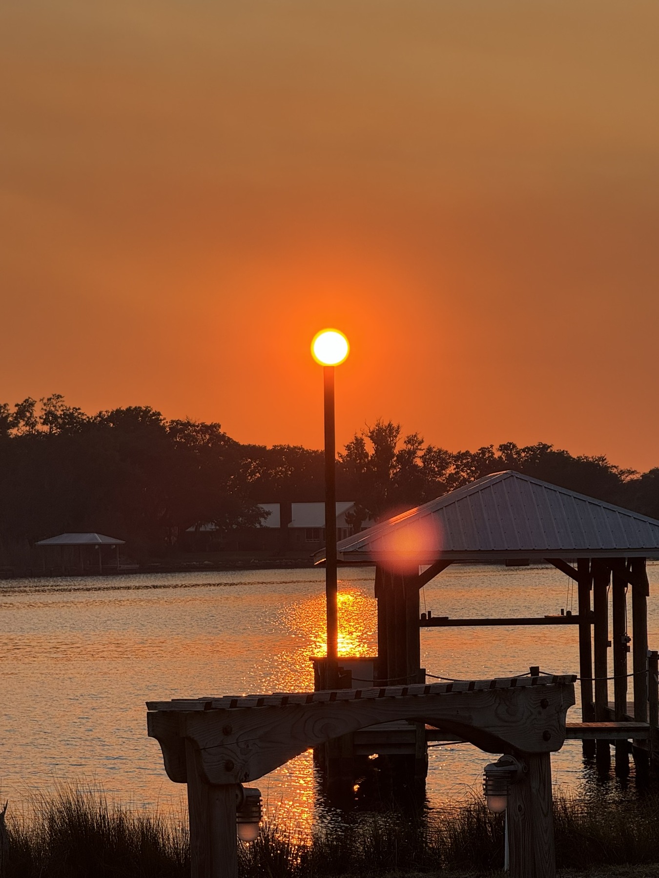 An orange sun aligns with a dock post beyond a pavilion